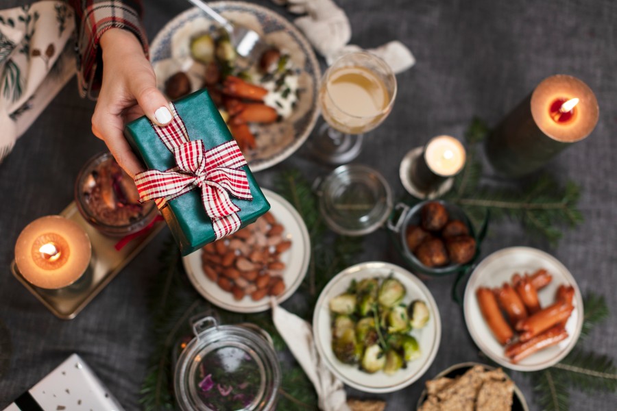 A hand holds a Christmas present in a green wrapper and a red-white blow over a table of food